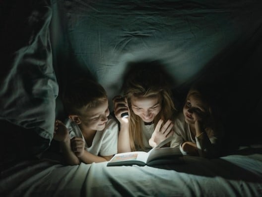 Two kids with mom reading with flashlight under blanket