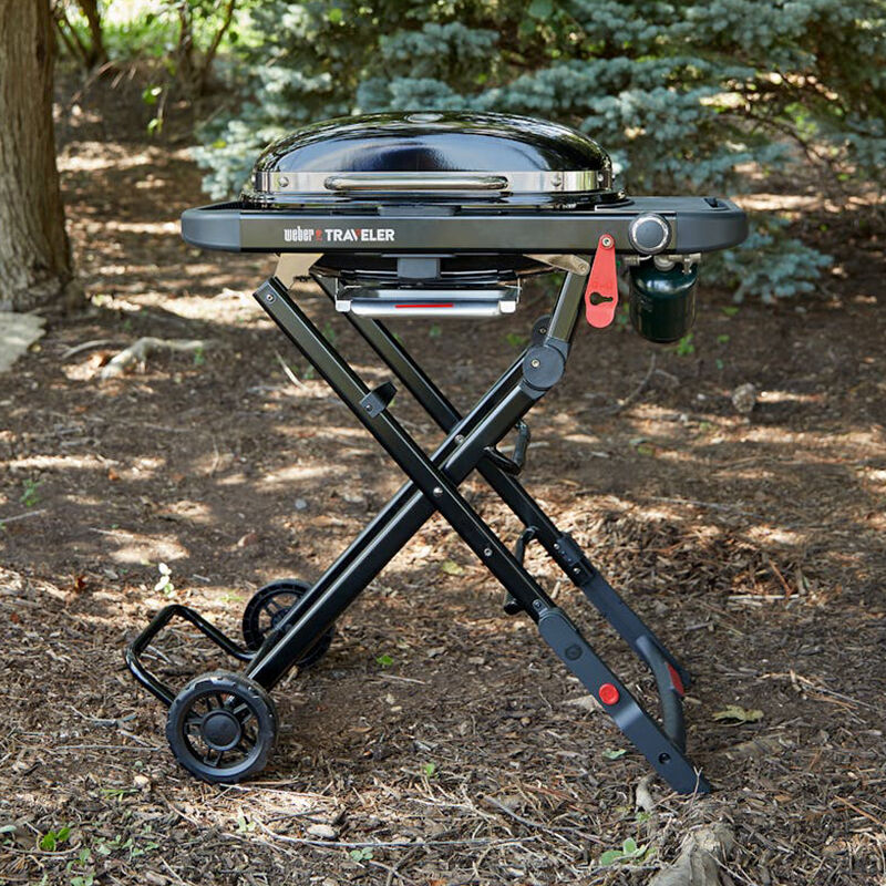 Black Weber Traveler portable gas grill on folding cart with wheels, set up outdoors on a wooded forest floor.
