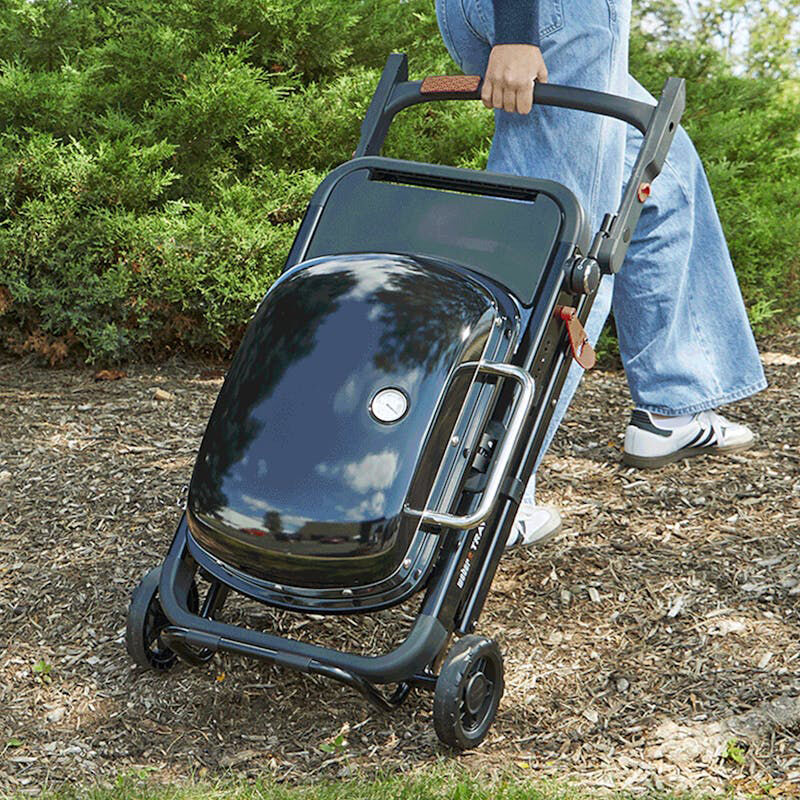 Person rolling a folded black Weber Traveler portable gas grill on wheels across a mulched outdoor path.