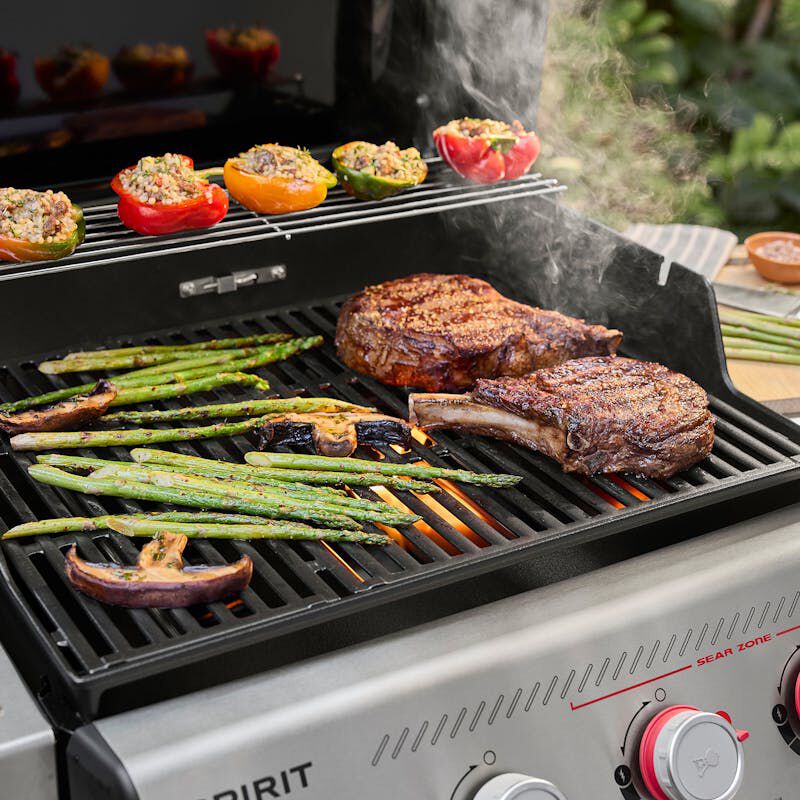 Weber Spirit E-425C cooking two thick ribeye steaks with asparagus and grilled vegetables on the main grate and stuffed peppers on the upper warming rack.