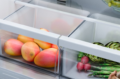 Close-up of Midea refrigerator's clear crisper drawers filled with fresh produce, including ripe mangoes in one compartment and green asparagus, radishes, and artichokes in the adjacent vegetable drawer.
