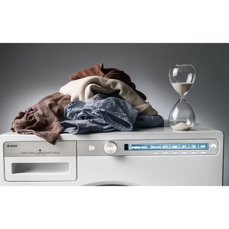 Top view of Asko washer control panel displaying digital interface and cycle options, with pile of jeans, towels, and hourglass arranged on top surface.