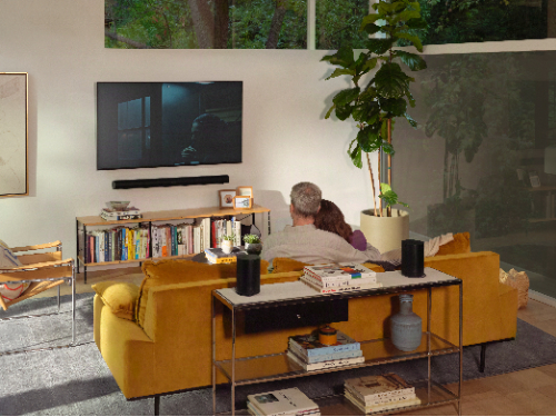 Lifestyle view of Sonos Era 100 SL speaker in black, displayed on an end table behind a couch where a couple is sitting while watching TV. 