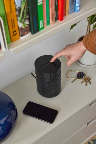 Sonos Era 100 SL speaker in black, shown plugged into a wall with a person's finger touching the top of the speaker.