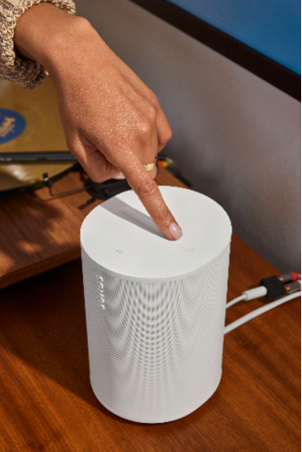 Sonos Era 100 SL speaker in white, shown plugged into a wall with a woman's finger touching the top of the speaker.