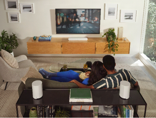 Lifestyle view of Sonos Era 100 SL speaker in white, displayed on an end table behind a couch where a couple is sitting while watching TV. 