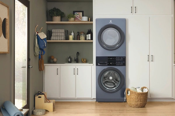 Electrolux titanium stacked washer and dryer installed in bright laundry room with white cabinets, open shelving, and modern decor including laundry baskets.