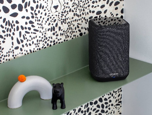 Denon Home 150 wireless speaker model HOME150BLACK in black placed on a green shelf against a patterned wall, shown in a modern decorative home setting.