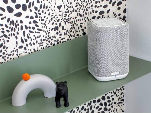 Denon Home 150 wireless speaker model HOME150WHITE in white placed on a green shelf against a patterned wall, shown in a modern decorative home setting.