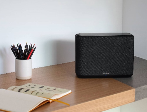 Denon Home 250 wireless speaker model HOME250BLACK in black placed on a modern wooden desk beside a notebook and pencil holder in a minimal home office setting.