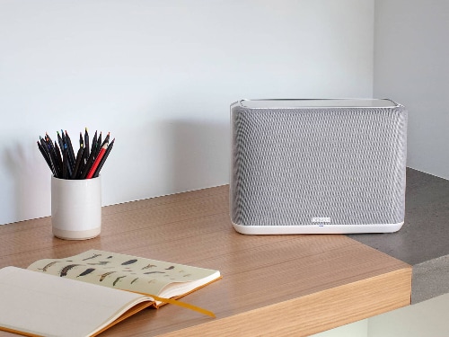 Denon Home 250 wireless speaker model HOME250WHITE in white placed on a modern wooden desk beside a notebook and pencil holder in a minimal home office setting.