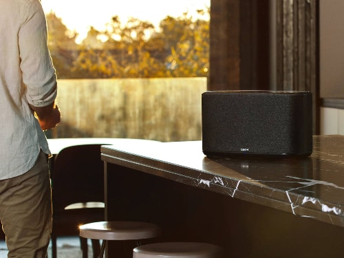 Denon Home 350 wireless speaker model HOME350BLACK in black placed on a marble counter in a sunlit modern kitchen with a person standing nearby.