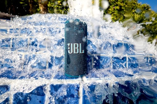 Blue JBL Grip portable Bluetooth speaker standing on blue tile under splashing water, demonstrating its waterproof design.