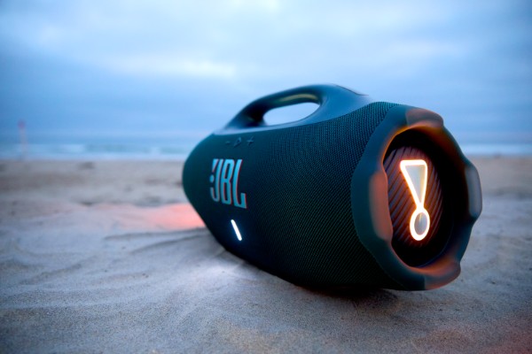 Black JBL Boombox 4 portable Bluetooth speaker resting on sand at the beach during dusk, with its side light glowing and ocean waves visible in the background