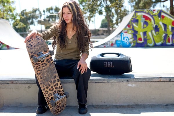 Black JBL Boombox 4 portable speaker on a skate park ledge beside a person holding a leopard-print skateboard, with colorful graffiti and ramps in the background.