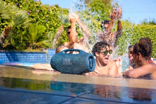 Blue JBL Boombox 4 portable Bluetooth speaker placed poolside while a group of people splash and enjoy a sunny pool party surrounded by greenery.