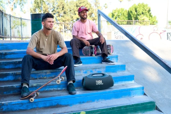 Blue JBL Boombox 4 portable speaker resting on skate park steps beside two people with skateboards, sitting and talking in the sun near ramps and graffiti.