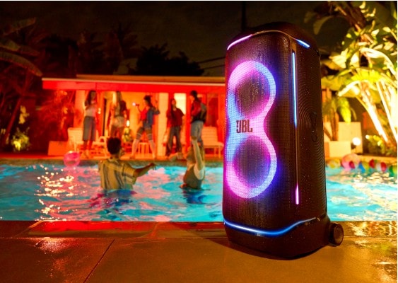 JBL PartyBox 720 speaker with glowing LED lights beside a pool at night, surrounded by people swimming and dancing at an outdoor party.