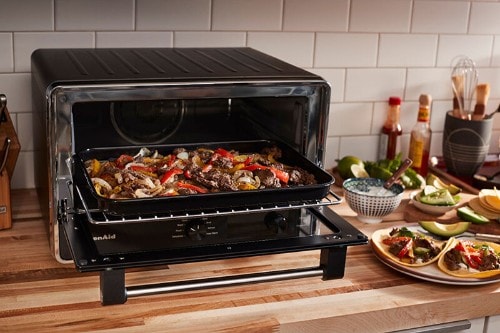 Black KitchenAid countertop toaster oven with glass door open, showing a sheet pan of roasted vegetables inside. The oven sits on a wooden counter with prepared