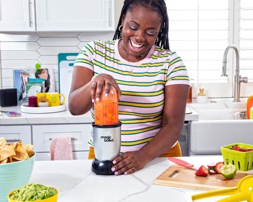 Smiling woman in a striped shirt blending a red smoothie with the Magic Bullet MBR-1101 blender in a bright kitchen, surrounded by fresh fruit, guacamole, and chips on the counter.