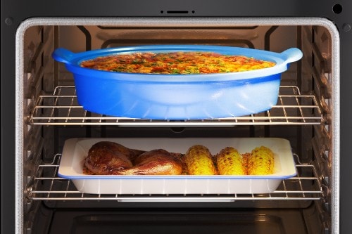 Midea oven interior with a blue casserole dish on the top rack and a tray of roasted chicken and corn on the bottom rack.