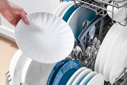 Hand placing a white plate into the lower rack of a Midea 18-inch dishwasher loaded with plates and silverware.