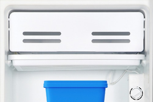 Close-up of compact refrigerator freezer compartment with white door featuring ventilation slots, showing a blue storage container and temperature control dial.