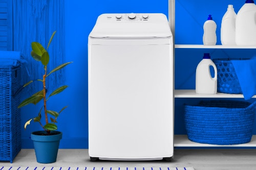 Midea top load washer in modern laundry room with blue walls, white shelving holding detergents, and blue plant pot, showcasing compact space-saving design.