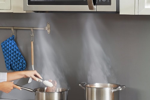 Hand holding spatula with steam rising from pots on stovetop, demonstrating Midea over-the-range microwave's ventilation system clearing cooking vapors in modern kitchen.