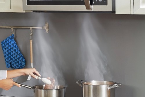 Midea over-the-range microwave venting steam upward from two cooking pots on a stovetop while food is being prepared.