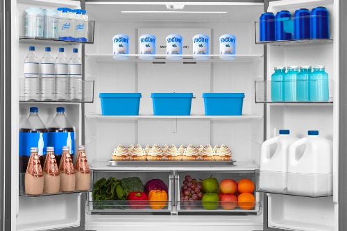 Interior view of Midea MRF23I5AST refrigerator stocked with yogurt, water bottles, blue containers, milk jugs, cupcakes, and fresh fruits and vegetables in crisper drawers.