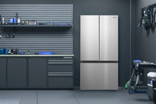 Midea MRF23I5AST stainless steel French door refrigerator standing in a modern garage workspace alongside dark tool cabinets, wall-mounted shelving, and organized tools.