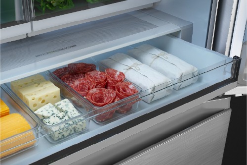 Close-up of Midea refrigerator pull-out deli drawer containing organized compartments of sliced salami, blue cheese, yellow cheddar cheese, and neatly wrapped white packages of deli meats.