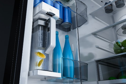 Close-up of Midea refrigerator interior AutoFill water pitcher with lemon slices, alongside blue glass bottles stored on the refrigerator door shelf.