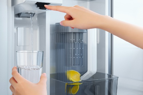 Person pressing interior water dispenser button on Midea refrigerator while holding a glass being filled with water, with lemon-infused AutoFill pitcher visible alongside.