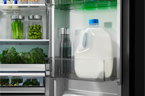 Interior view of Midea refrigerator door shelf holding a gallon of milk and a dark water bottle, with shelves in background storing fresh vegetables and glass jars of produce.