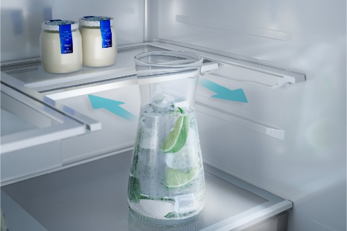 Close-up of Midea refrigerator interior showing a glass pitcher filled with ice water and lime slices on an adjustable shelf, with blue arrows indicating sliding shelf feature and yogurt containers visible above.