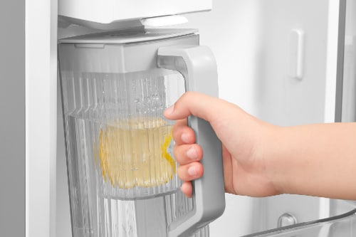 Hand holding clear pitcher with yellow beverage under AutoFill water dispenser inside refrigerator door compartment, showing automatic filling feature with water flowing into pitcher.