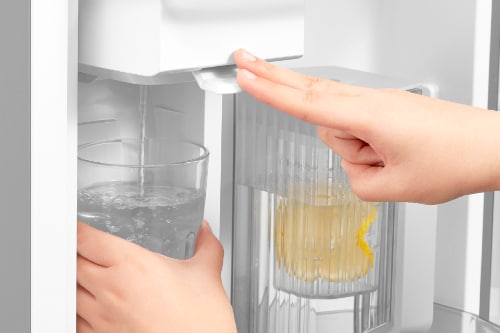 Two hands interacting with refrigerator dispenser - left hand holding cup under water spout while right hand presses button. Pitcher with yellow beverage visible in door shelf behind.