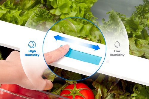 Close-up of the Midea 18 cu. ft. Top-Freezer Refrigerator model MRT18D3B showing a hand adjusting the crisper drawer's humidity control slider, with fresh lettuce and tomatoes inside.