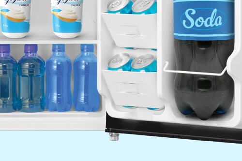 Close-up of Midea mini fridge's adjustable leveling leg at bottom corner with interior shelves visible showing yogurt, water bottles, beverage cans, and two-liter soda bottle in door.