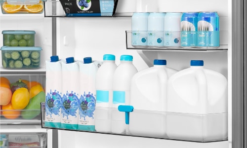 Close-up of Midea refrigerator door bins showing gallon milk jugs, water bottles, and beverage cans organized in deep storage compartments with fresh produce visible on shelves.