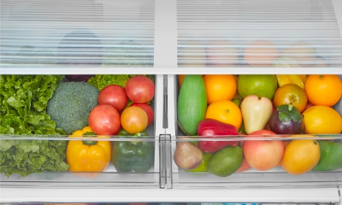 Midea refrigerator's dual humidity-controlled crisper drawers filled with colorful fresh produce including lettuce, broccoli, tomatoes, peppers, squash, and citrus fruits for optimal freshness.