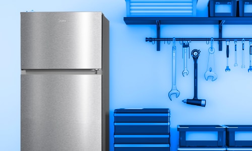 Midea stainless steel top-freezer refrigerator positioned in garage workshop setting with blue tool storage and hanging wrenches, demonstrating garage-ready capability.