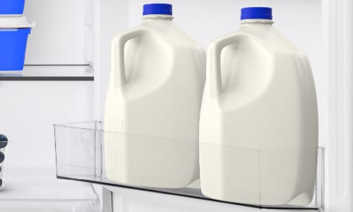 [Close-up of Midea refrigerator door shelf holding two gallon milk jugs with blue caps, demonstrating spacious door storage capacity.