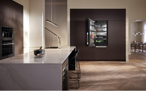 Modern luxury kitchen featuring Miele left-hinged built-in refrigerator with flush-mounted design in dark brown cabinetry, illuminated interior visible through open doors, complemented by white marble waterfall island, designer bar stools, and contemporary pendant lighting.