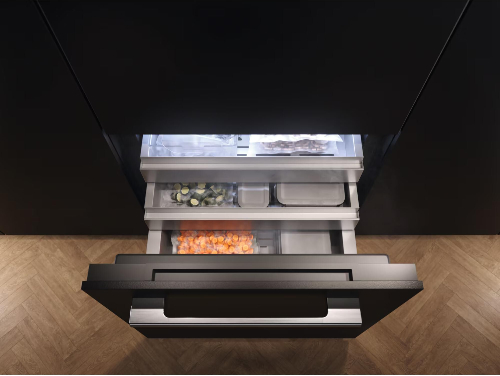 Miele refrigerator bottom freezer drawer fully extended showing tri-level organization system with ice cube tray on top shelf, frozen vegetables in middle compartment, and pizza and frozen items in lower drawer, featuring LED lighting and black exterior finish.