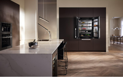 Modern luxury kitchen featuring Miele built-in French door refrigerator with illuminated interior in dark brown cabinetry, complemented by white marble waterfall island, designer bar stools, contemporary pendant lighting, and open floor plan to dining area.