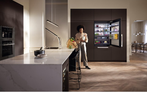 Modern luxury kitchen with woman holding flowers at white marble waterfall island, Miele built-in refrigerator with illuminated interior in dark brown cabinetry, designer bar stools, and contemporary pendant lighting creating elegant living space.