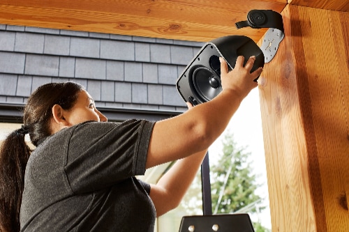 Person installing a black Sonos Outdoor Speaker model OUTDRWW1BLK on a wooden beam under an exterior roof, showing the durable, weather-resistant mount and outdoor setup.
